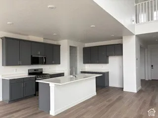 Kitchen featuring dark charcoal cabinetry, white stone-finish countertops, a central island with an integrated sink, built-in stainless steel appliances, and wood-finish flooring