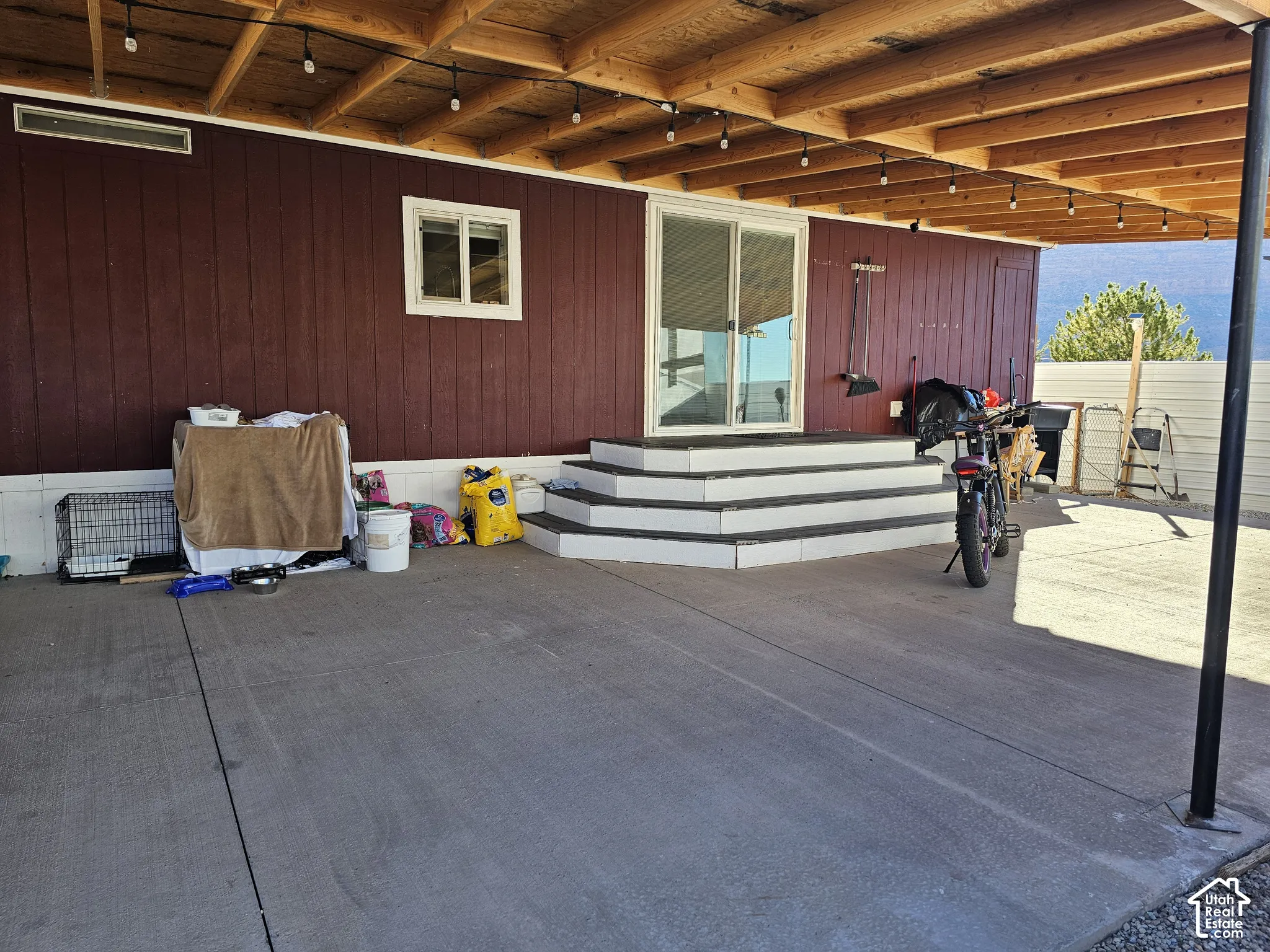 Covered concrete patio featuring an exposed timber beam ceiling, sliding glass entry door, and tiered exterior stairs