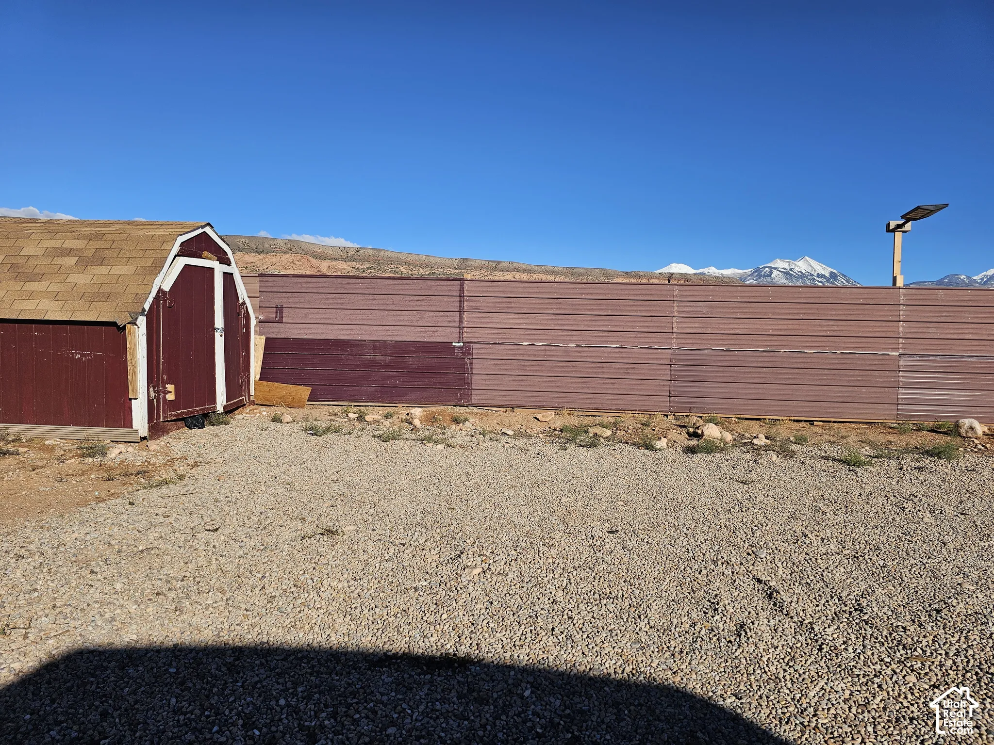 Exterior storage structure with a shingle roof, enclosed by a corrugated metal fence on a gravel lot, featuring a backdrop of snow-capped mountains