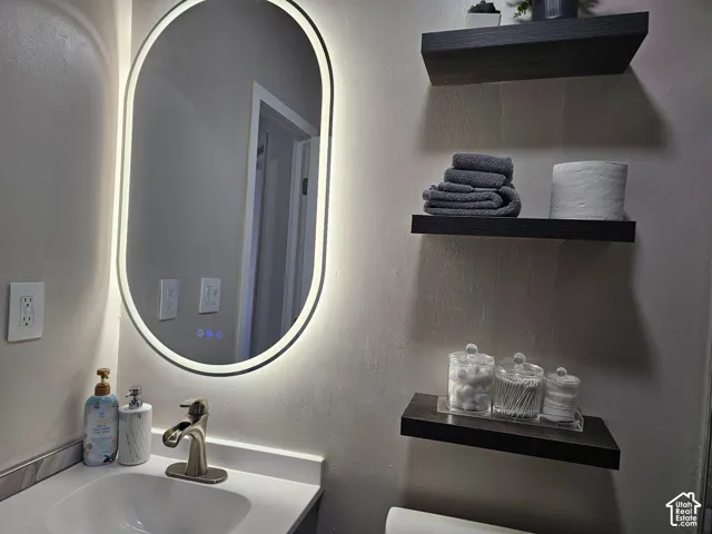 Oval-shaped LED-backlit mirror, integrated single-handle faucet, solid-surface vanity countertop, and three wall-mounted dark-finish wood shelves
