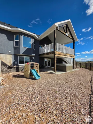 Rear exterior featuring a covered elevated deck with timber beams and a metal railing, a ground-level concrete patio, and a gravel-surfaced yard enclosed by metal fencing