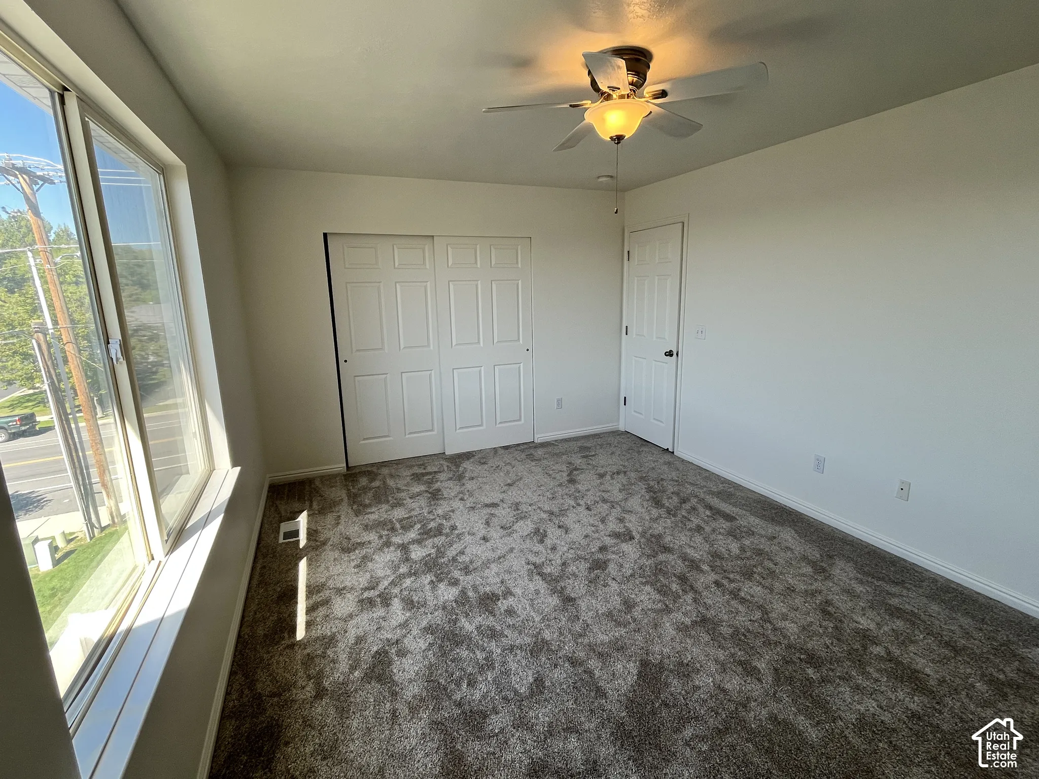 Neutral-toned carpeted room featuring a large picture window, sliding-panel closet, white interior door, and a ceiling fan with integrated lighting