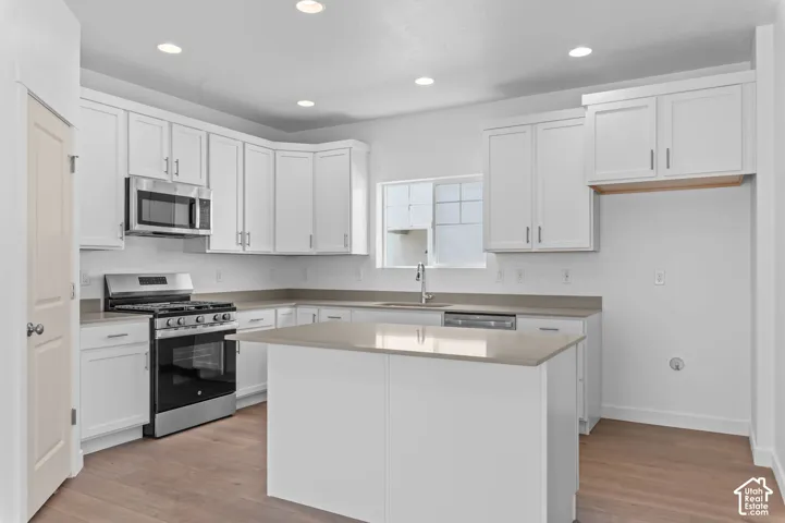 Kitchen featuring white shaker-style cabinetry, neutral-toned solid surface countertops, stainless steel appliances, a central island, and wood-finish flooring