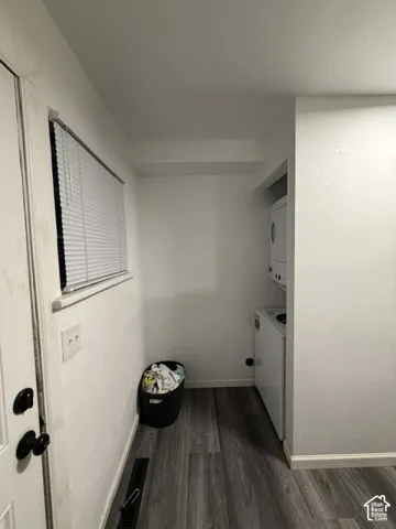 Laundry area featuring wood-finish flooring, a stacked washer and dryer unit, and a window equipped with horizontal blinds