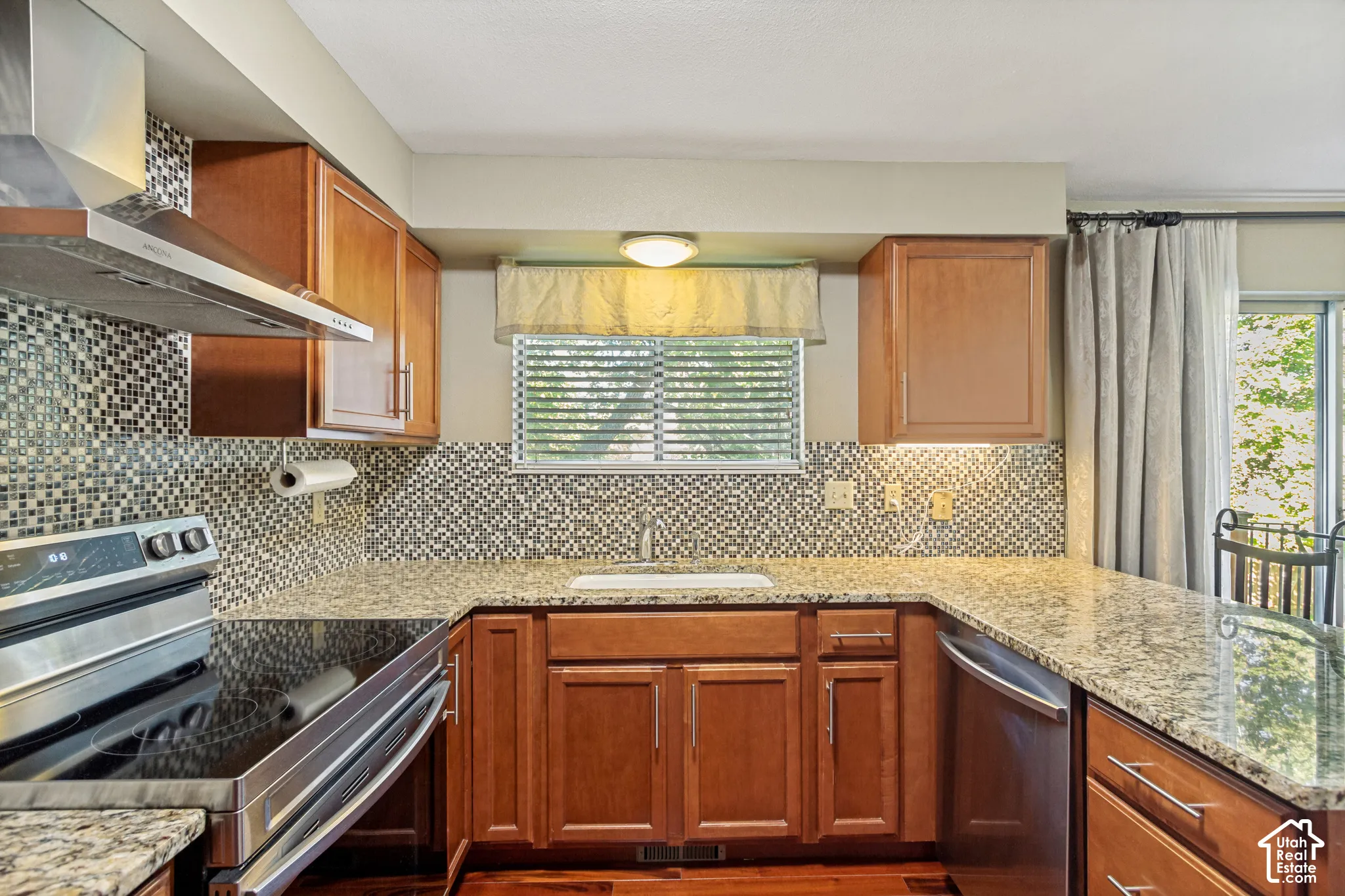 Kitchen featuring wood cabinetry, stone-finish countertops, decorative mosaic tile backsplash, stainless steel range hood, and under-cabinet lighting