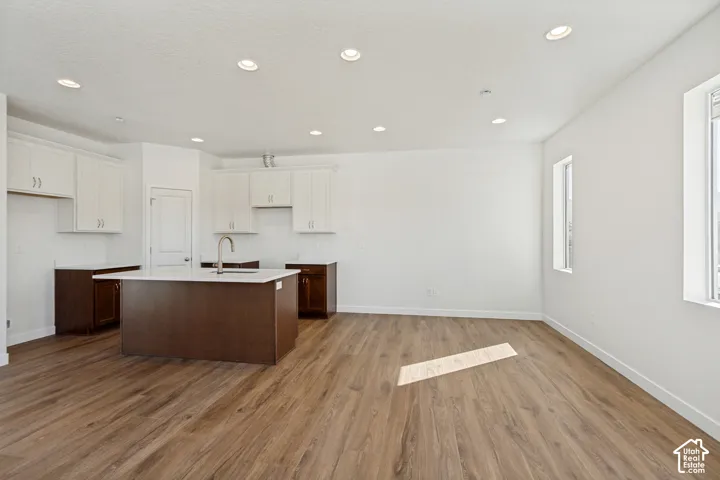 Kitchen featuring wood-finish flooring, recessed lighting, white upper cabinetry, dark base cabinetry, and a central island with a solid-surface countertop and faucet