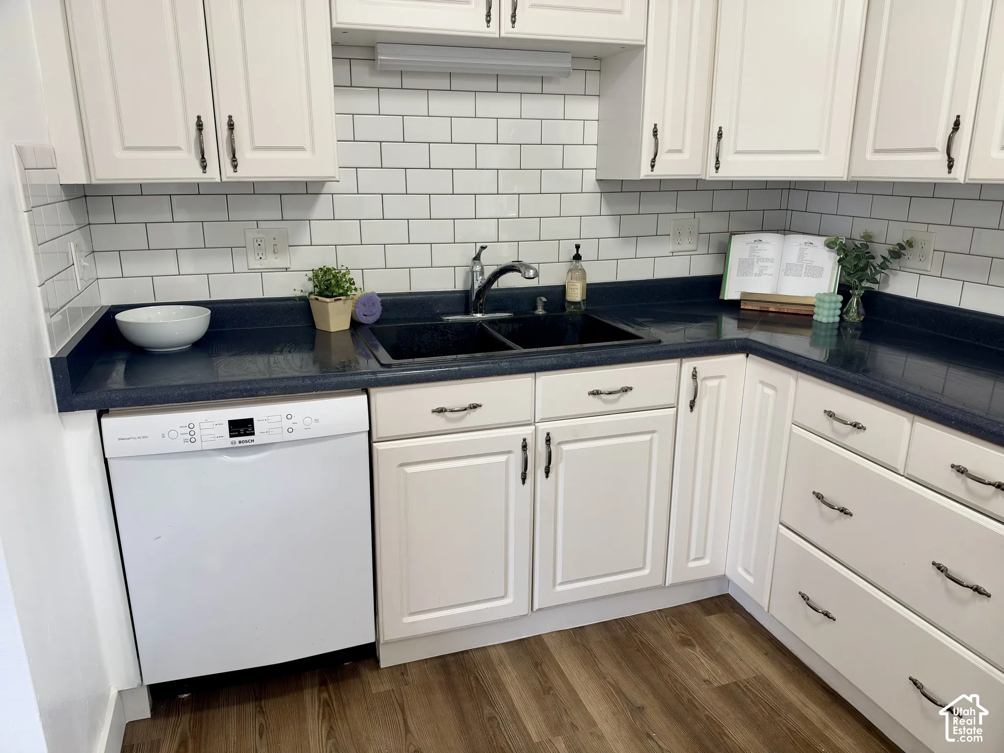 Kitchen featuring white cabinetry, dark countertops, white subway tile backsplash, dual-basin sink with pull-down faucet, and built-in dishwasher