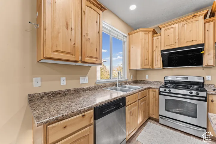 Kitchen featuring natural-finish wood cabinetry, stone-pattern laminate countertops, stainless steel dual-basin sink with arched faucet, stainless steel dishwasher, and built-in microwave and gas range
