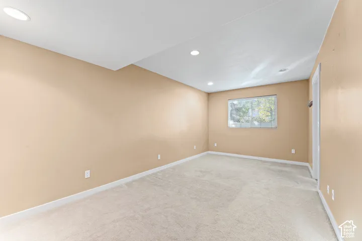 Neutral-toned interior space featuring light-colored wall-to-wall carpeting, recessed ceiling lighting, and a single window with blinds
