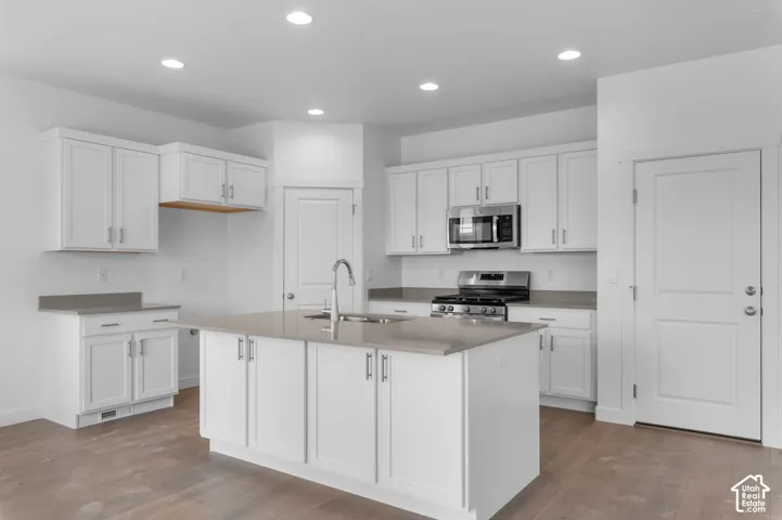 Kitchen featuring white shaker-style cabinetry, quartz countertops, stainless steel appliances, a central island with an undermount sink, and wood-finish flooring