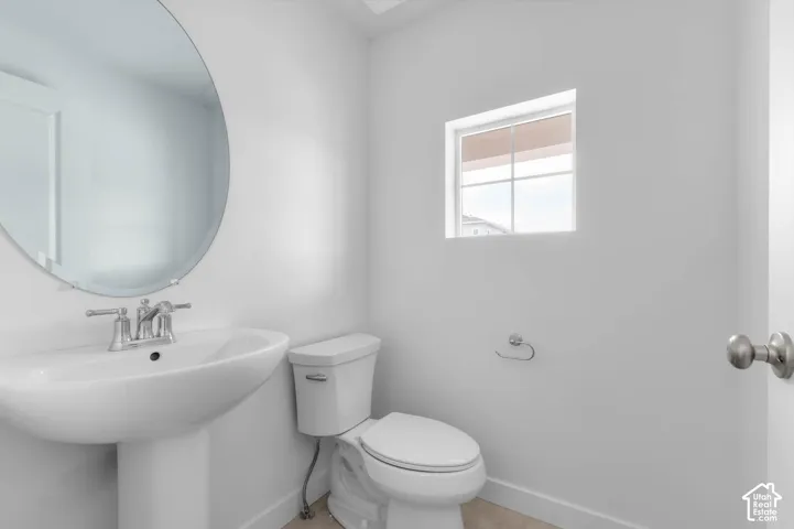 Pedestal sink featuring a polished chrome faucet, round wall-mounted mirror, white ceramic toilet, and a window with a grid pattern