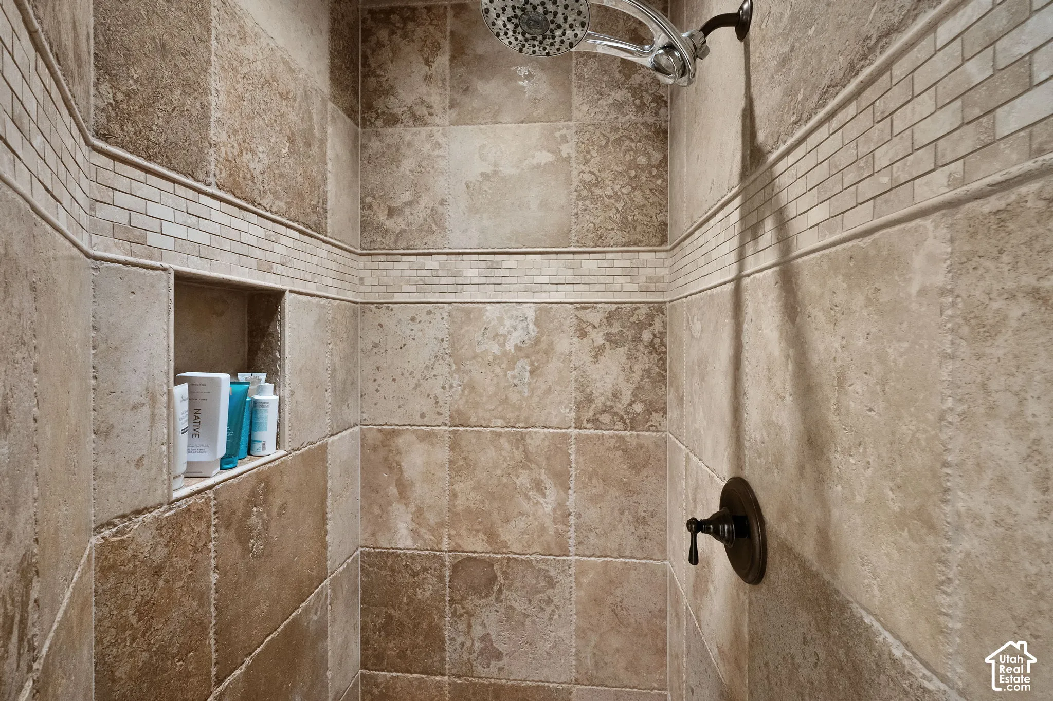Walk-in shower featuring natural stone wall tile, custom tile inlay accent border, integrated recessed shelving, and a wall-mounted shower head with oil-rubbed bronze fixtures