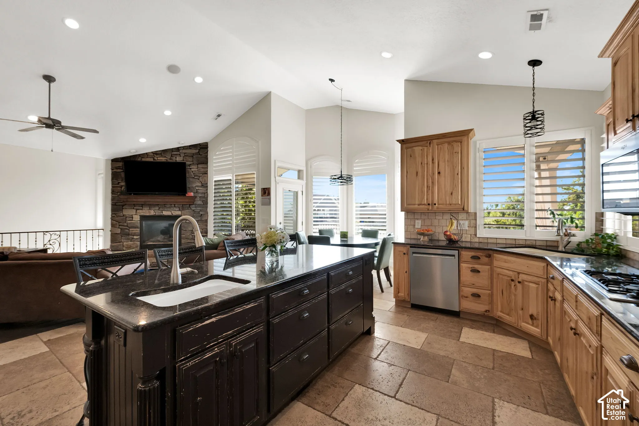 Kitchen featuring natural-finish wood cabinetry, a dark-toned center island with an integrated farmhouse sink, stone tile flooring, recessed lighting, and a floor-to-ceiling stone fireplace surround