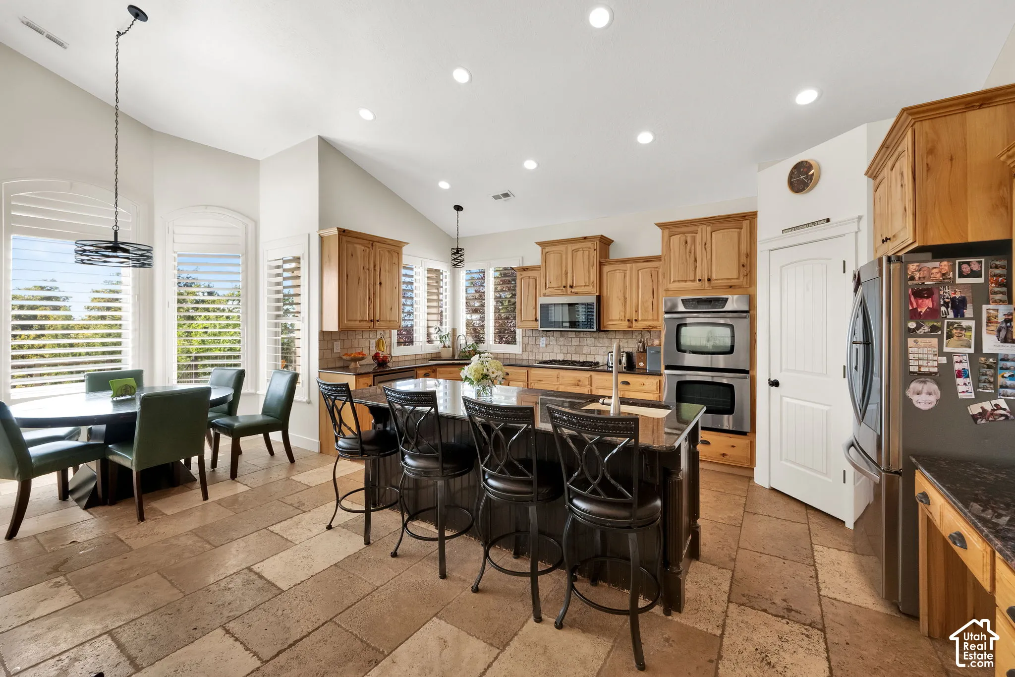 Kitchen featuring natural wood cabinetry, stone-tiled flooring, a dark-toned center island with a stone countertop, built-in double wall ovens, and arched windows with plantation shutters