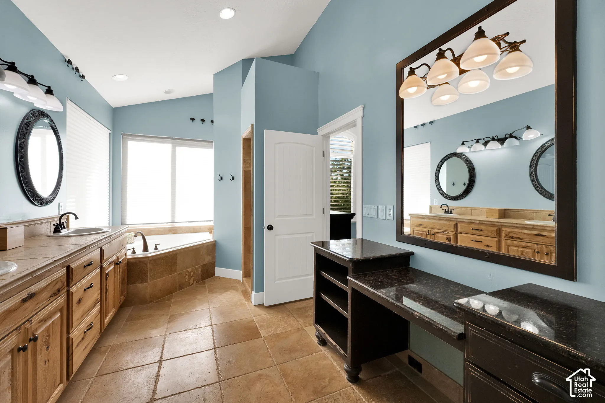 Bathroom featuring a dual-sink vanity with wood cabinetry, stone-tiled soaking tub surround, stone tile flooring, a dark-finished built-in vanity station, and large framed wall mirrors
