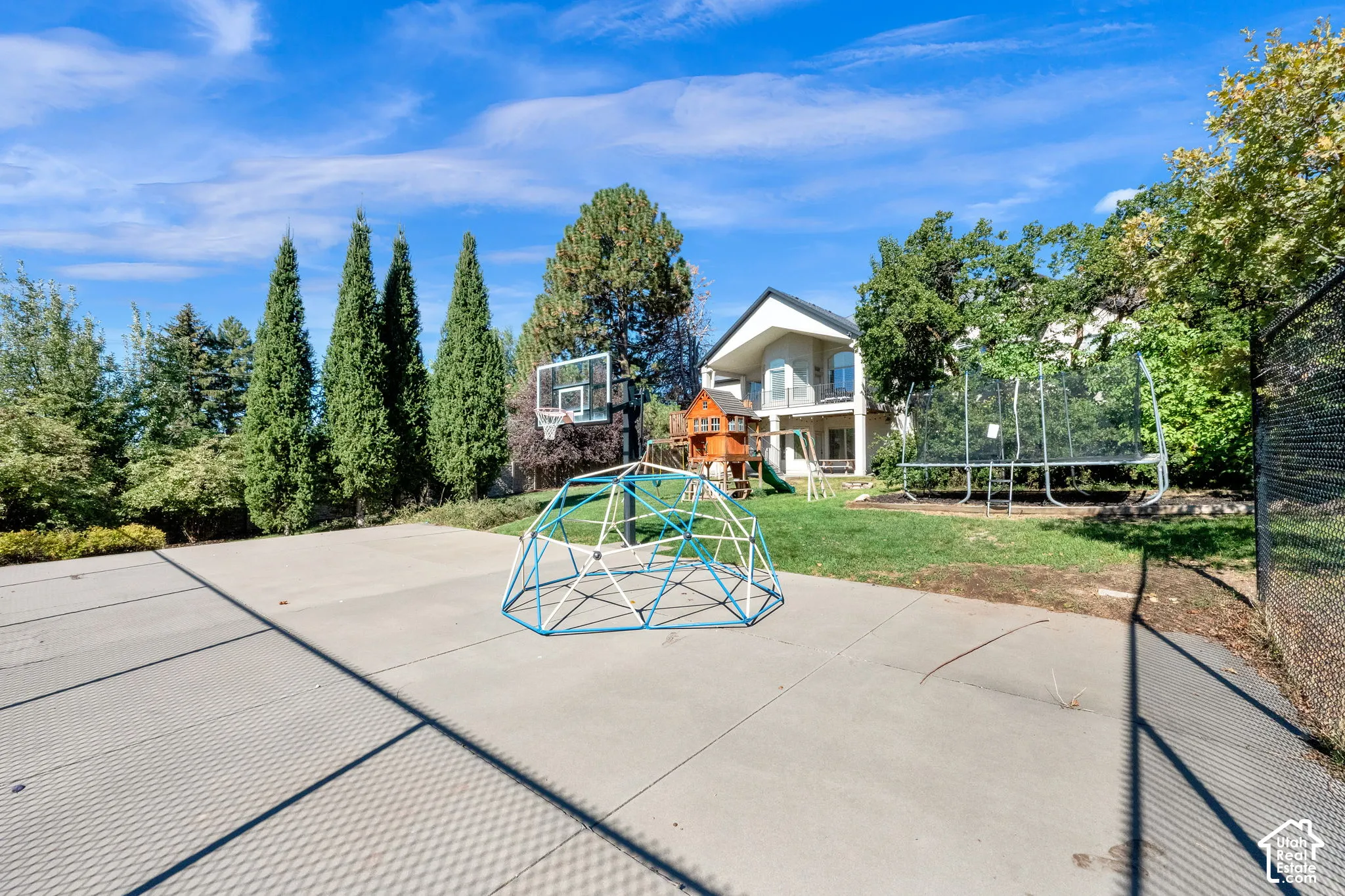 Backyard featuring a concrete court, chain-link fencing, mature trees, and a landscaped lawn