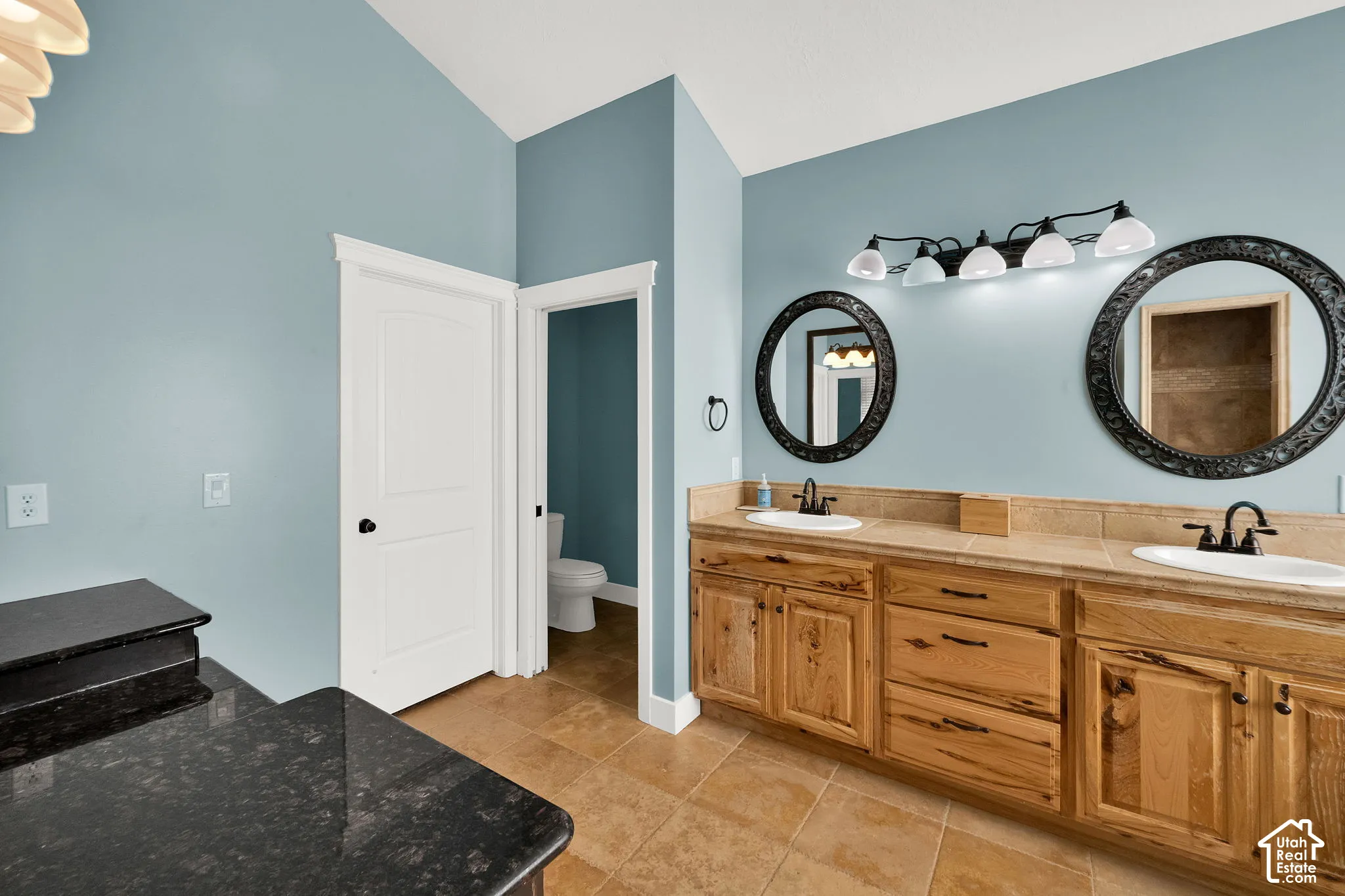 Dual-sink vanity with natural wood cabinetry, stone-style countertop, dual circular mirrors, and multi-light wall fixture