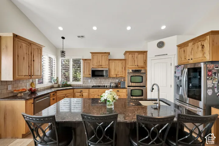 Kitchen featuring natural wood cabinetry, stone countertops, a central island with integrated sink, stainless steel double wall ovens, and recessed lighting