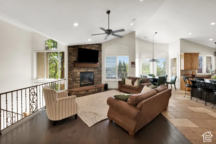 Open-concept living space featuring vaulted ceilings, floor-to-ceiling stone-clad fireplace, iron railing, recessed lighting, and a blend of dark wood-finish flooring and neutral-tone stone flooring