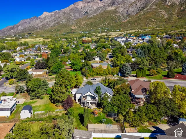 Aerial perspective featuring a multi-level residence with a gray shingle roof, integrated upper-level deck, fenced backyard, and private sports court, all set against a prominent mountain backdrop