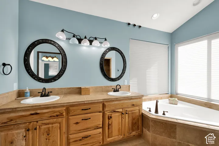 Dual-sink vanity with wood cabinetry, tile countertops, round wall-mounted mirrors, and built-in soaking tub with tile surround