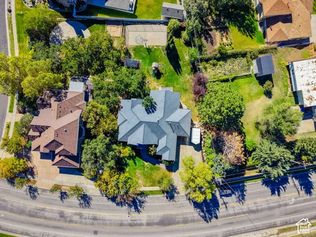 Aerial perspective featuring a multi-gabled roofline, expansive grass yard, mature perimeter landscaping, concrete driveway, and a paved hardscape area