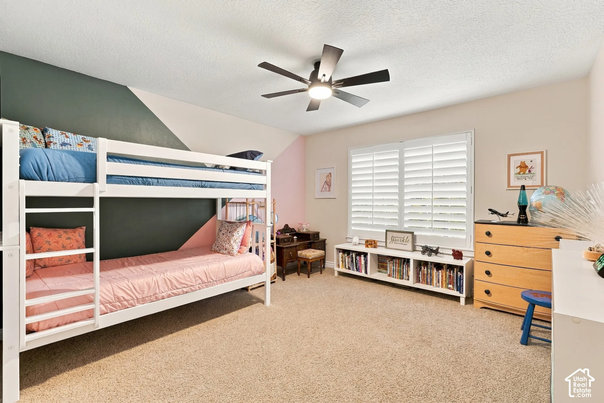 Bedroom featuring carpeted flooring, neutral wall paint with geometric accent wall, white plantation shutters, and a central ceiling fan with integrated lighting