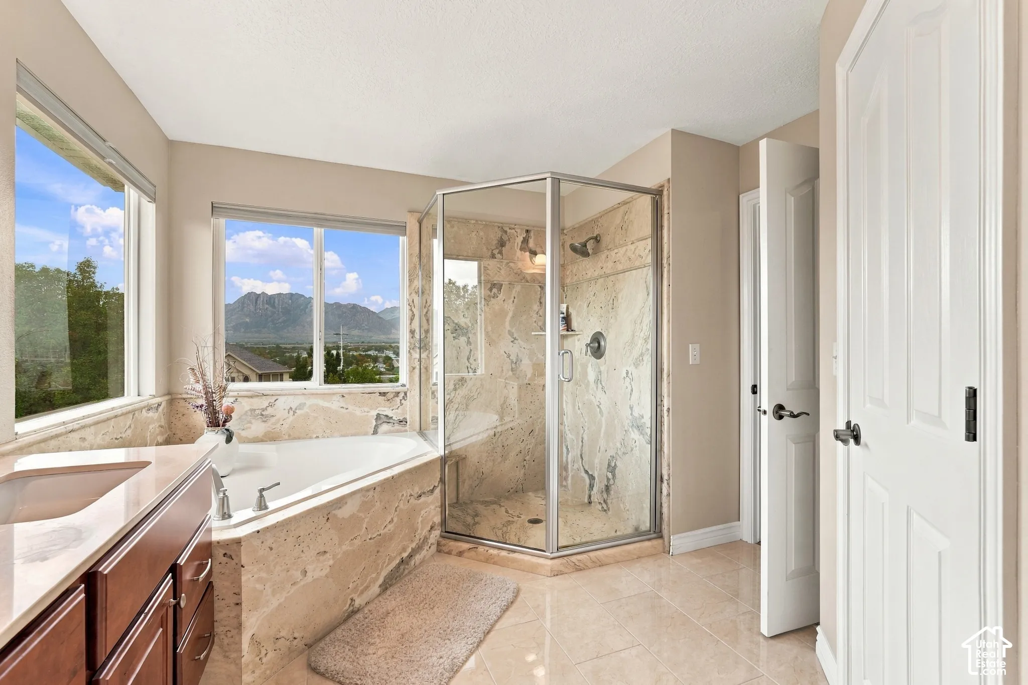 Corner soaking tub and separate glass-enclosed shower featuring stone tile surround, stone-topped vanity, and mountain scenery