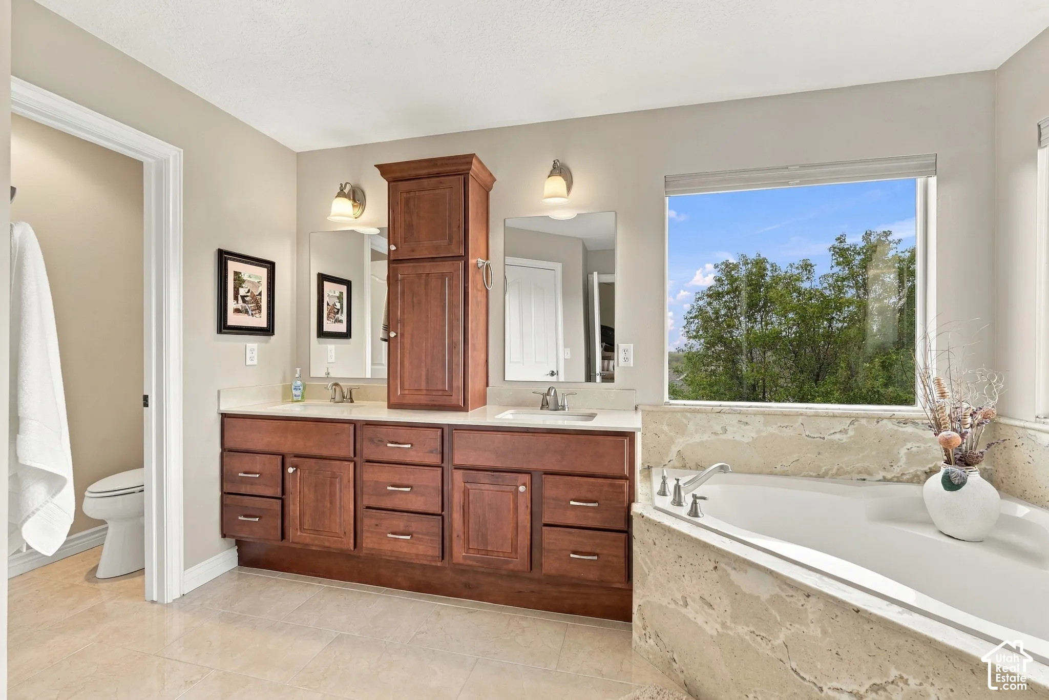 Dual-sink vanity with wood-finish cabinetry, stone countertop, central storage tower, and built-in soaking tub with stone surround