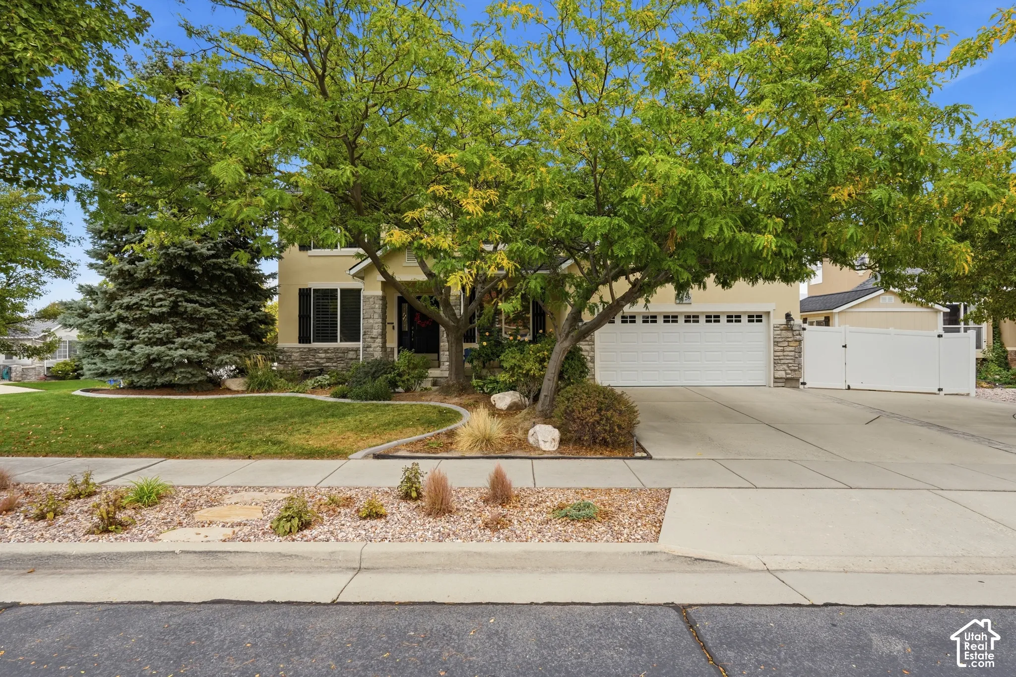 Two-story residence featuring a stone-accented facade, attached multi-car garage, concrete driveway, and xeriscaped front landscape with mature trees