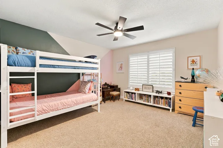 Bedroom featuring carpeted flooring, neutral wall paint with geometric accent wall, white plantation shutters, and a central ceiling fan with integrated lighting