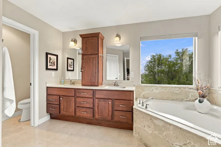 Dual-sink vanity with wood-finish cabinetry, stone countertop, central storage tower, and built-in soaking tub with stone surround