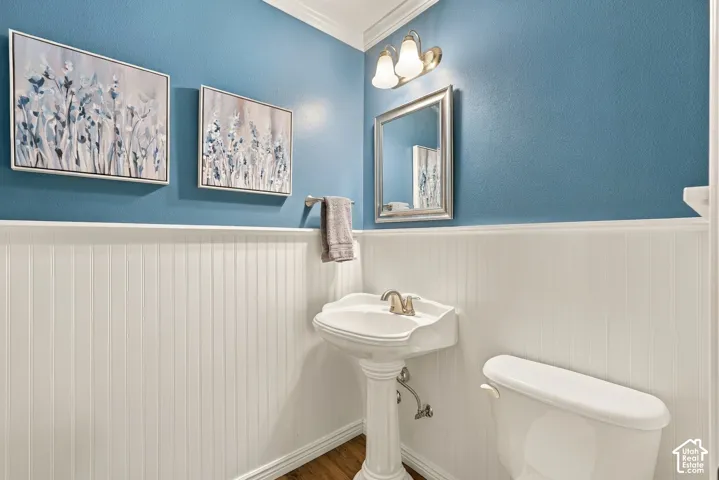 White beadboard wainscoting, pedestal sink with metallic fixtures, wall-mounted mirror, two-light wall sconce, and wood-finish flooring