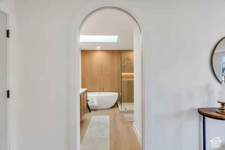 Arched entryway featuring wood-finish flooring, freestanding soaking tub, glass-enclosed shower, and vertical wood-slat accent wall with integrated skylight