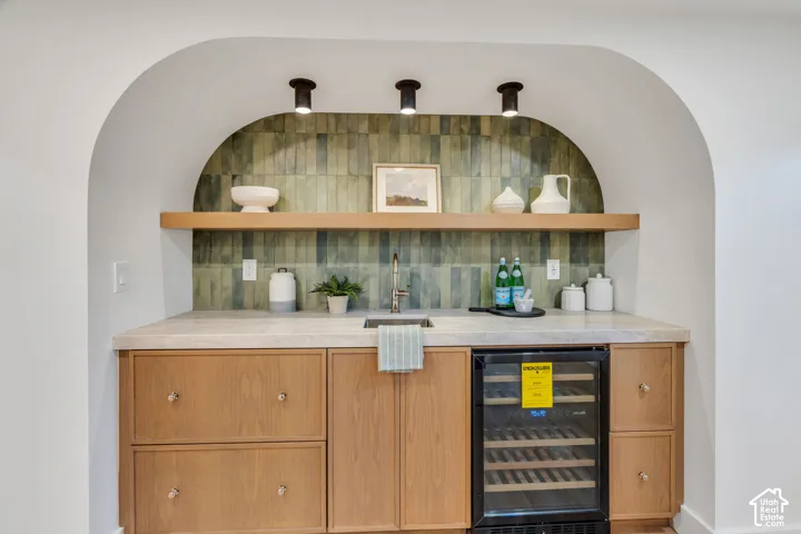 Built-in arched wet bar featuring wood cabinetry, natural stone countertop, vertical ceramic tile backsplash, integrated beverage cooler, and floating wood shelf with recessed overhead lighting