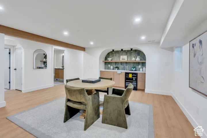 Custom arched wet bar featuring wood-finish cabinetry, a tiled backsplash, built-in beverage refrigerator, and open floating shelving, complemented by wood-finish flooring and recessed lighting throughout