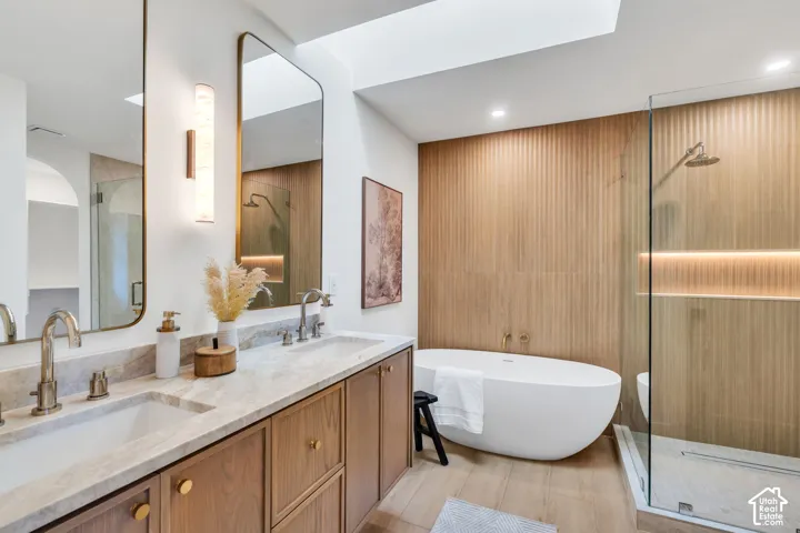 Dual-sink vanity with stone countertop and brass fixtures, freestanding oval soaking tub, floor-to-ceiling ribbed wood-finish accent wall, walk-in glass-enclosed shower with integrated niche lighting, and recessed skylight