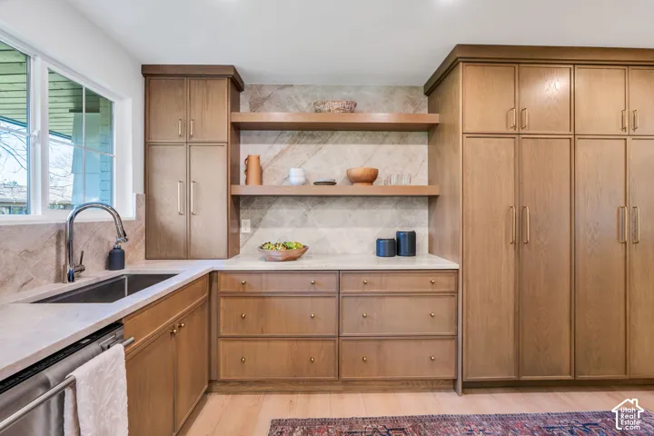 Kitchen featuring wood-finish cabinetry, stone slab backsplash, integrated shelving, stone countertops, and an undermount sink with a gooseneck faucet