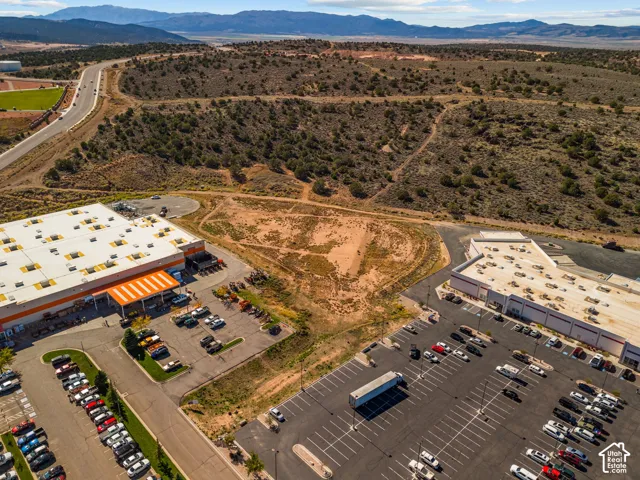 Expansive lot featuring terrain with native vegetation, positioned between developed commercial structures with asphalt parking lots, set against a mountainous horizon