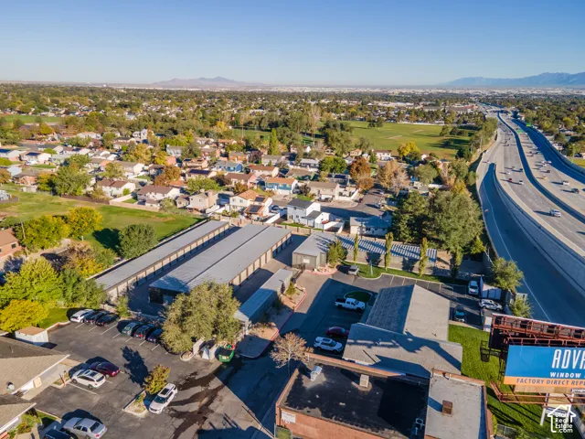 Aerial perspective featuring a multi-unit storage facility with metal roofing, asphalt-paved parking areas, and mature trees situated near a highway