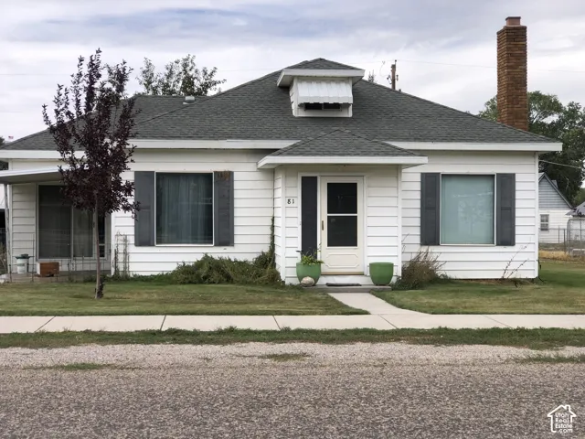 Single-story exterior featuring white horizontal siding, dark window shutters, an asphalt shingle roof, and a prominent brick chimney