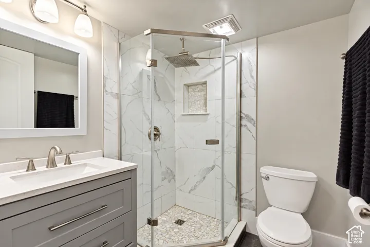 Bathroom featuring a gray vanity with stone countertop, corner glass-enclosed shower with tiled surround and stone-inlay niche, overhead rainfall showerhead, and a framed mirror with integrated lighting
