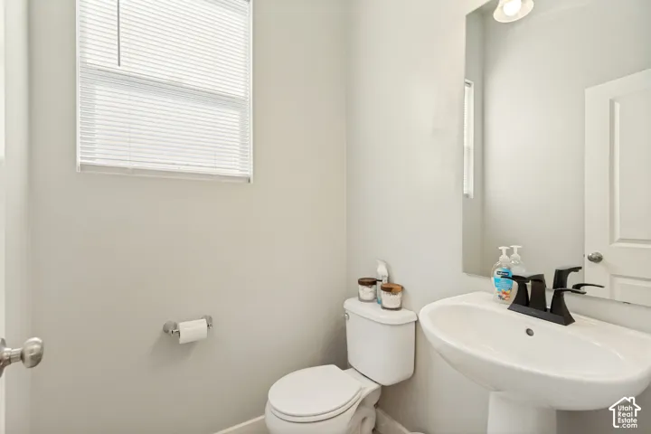Pedestal sink with a matte black faucet, wall-mounted mirror, white toilet, and window with integrated blinds