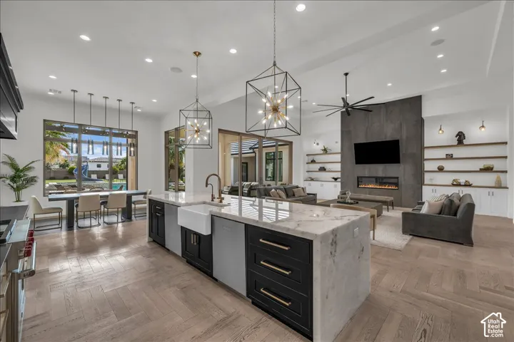 Open-concept living space featuring wood-finish herringbone flooring, a large kitchen island with a farmhouse sink and stone-surround countertop, ceiling-height fireplace wall with integrated shelving, and geometric pendant lighting