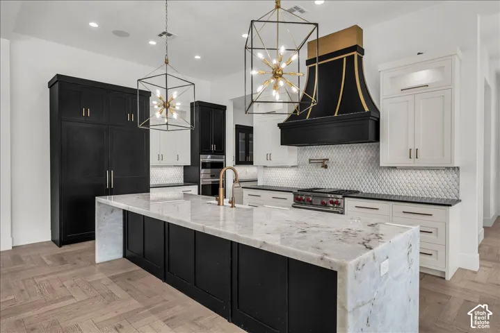 Kitchen featuring a large central island with waterfall-edge stone countertops, custom two-tone cabinetry, a bespoke range hood, a tiled backsplash, and geometric pendant lighting