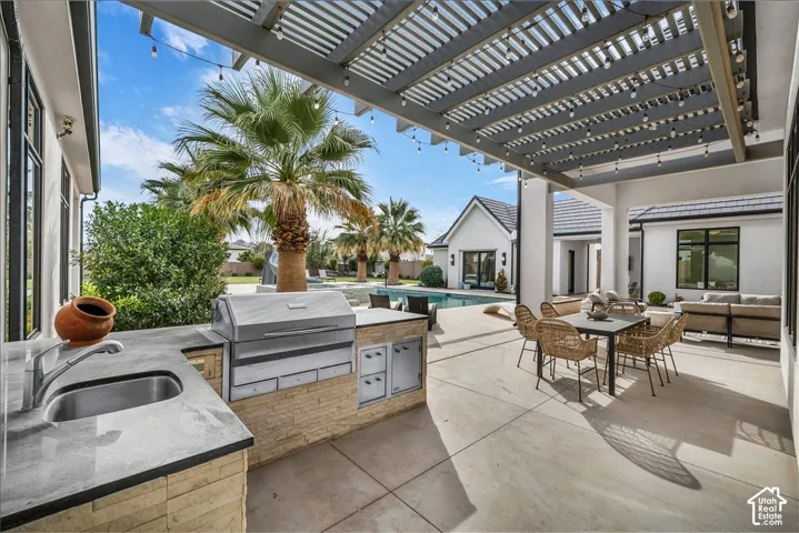 Covered patio featuring an outdoor kitchen with a built-in stainless steel grill and sink, stone-veneer cabinetry, concrete hardscaping, and a pergola structure