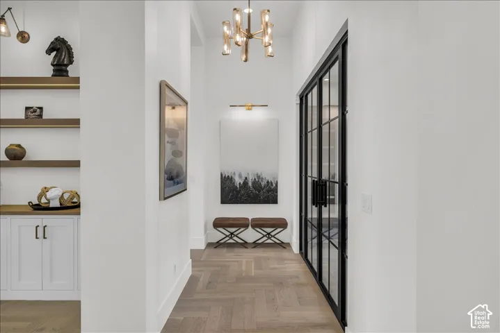 Herringbone-pattern wood-finish flooring, built-in cabinetry with integrated shelving, glass-paneled double doors with matte black frames, multi-arm chandelier, and wall-mounted vanity lighting
