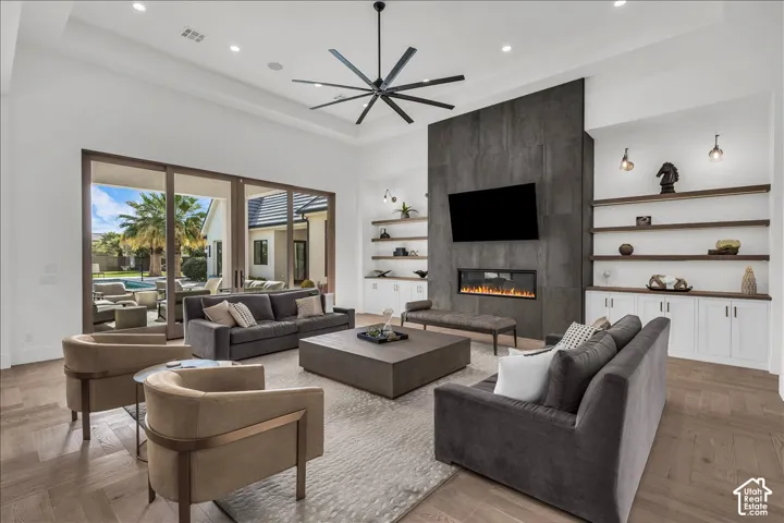 Great room featuring expansive tray ceilings, wood-finish flooring, a floor-to-ceiling charcoal stone fireplace, built-in cabinetry with wood shelving, and multi-panel sliding glass doors