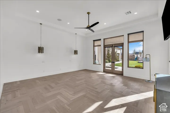 Herringbone-patterned wood-finish flooring, tray ceiling, dual metal pendant lighting, matte black ceiling fan, and double glass-paneled exterior doors with flanking picture windows