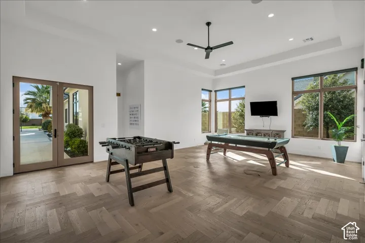 Spacious room featuring herringbone-pattern wood-finish flooring, tray ceilings, recessed lighting, and large windows with brown frames, complemented by dual glass-panel doors